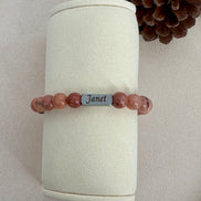 Beaded bracelet with a name tag on a cylindrical white stand with a pine cone in the background.