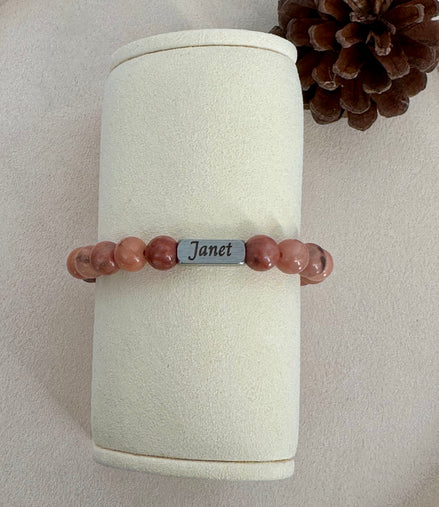 Beaded bracelet with a name tag on a cylindrical white stand with a pine cone in the background.