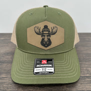 Green and beige trucker hat with moose  wearing a beanie and sunglasses that has been laser engraved on a leatherette patch on the front of the hat