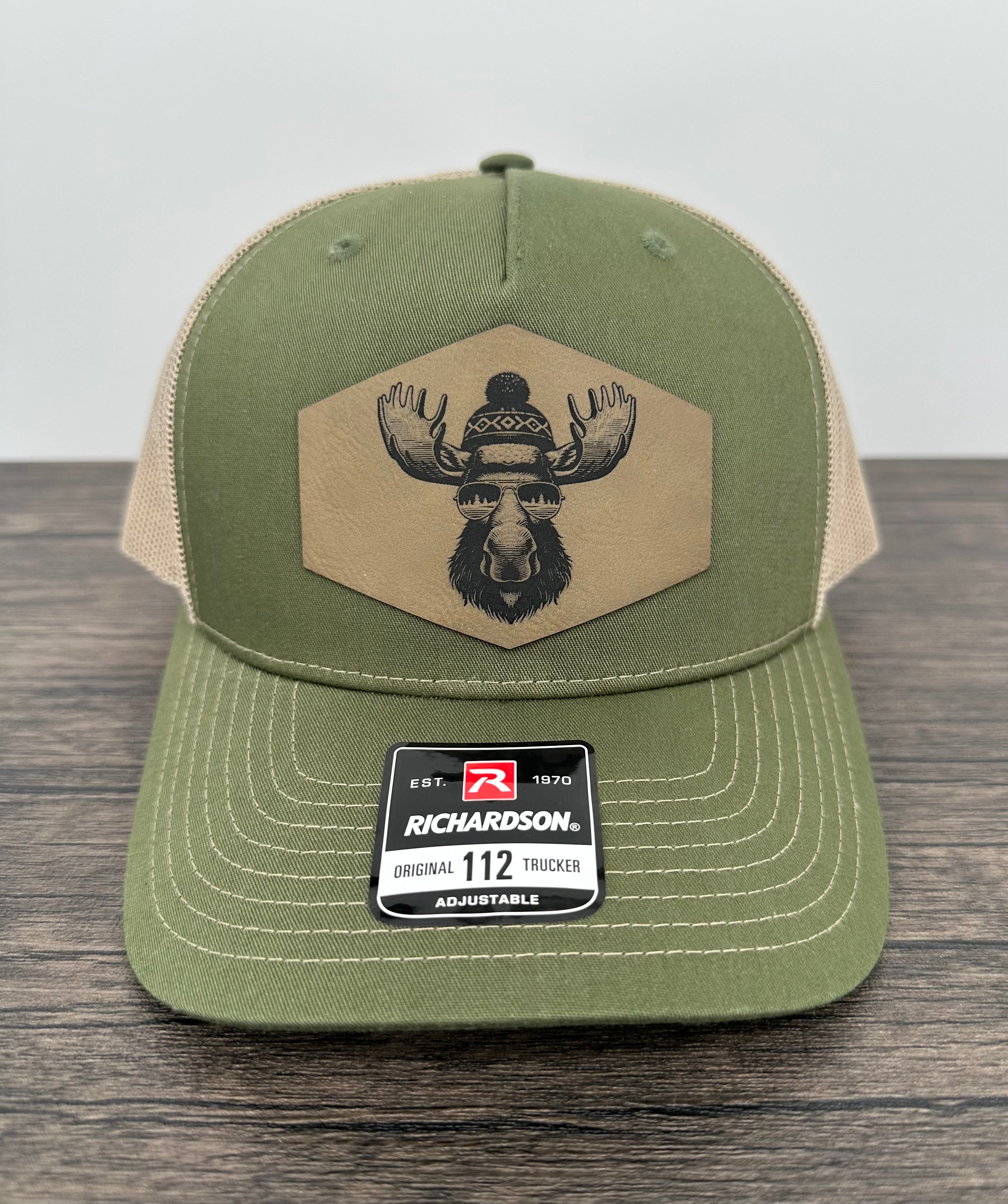 Green and beige trucker hat with moose  wearing a beanie and sunglasses that has been laser engraved on a leatherette patch on the front of the hat