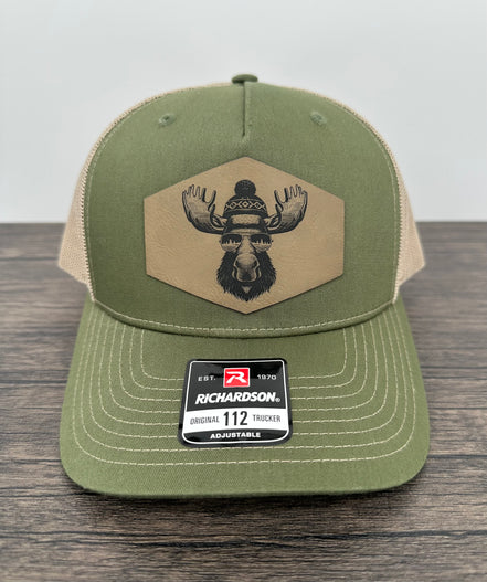 Green and beige trucker hat with moose  wearing a beanie and sunglasses that has been laser engraved on a leatherette patch on the front of the hat