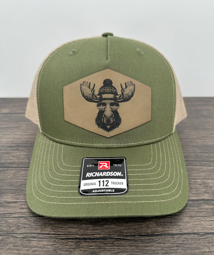 Green and beige trucker hat with moose  wearing a beanie and sunglasses that has been laser engraved on a leatherette patch on the front of the hat