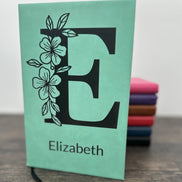 Custom Laser Engraved Notebook with floral design with a large letter and name engraved on the cover. The notebook is Seafoam green and you can see other colors in the background in pink, purple, brown, red, blue and black