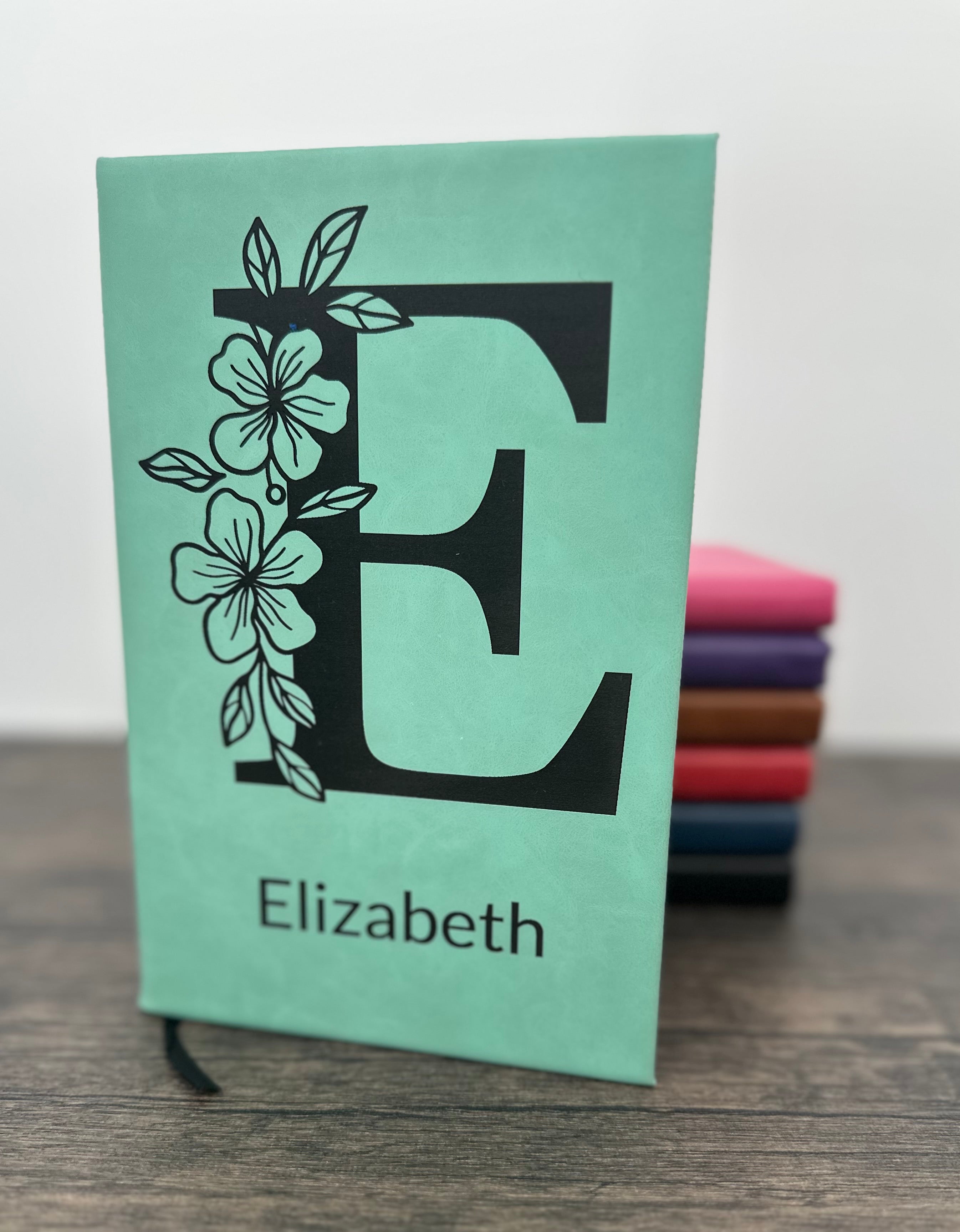 Custom Laser Engraved Notebook with floral design with a large letter and name engraved on the cover. The notebook is Seafoam green and you can see other colors in the background in pink, purple, brown, red, blue and black