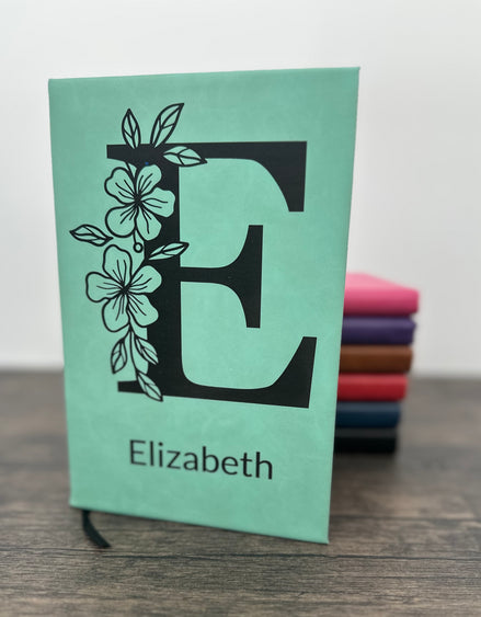 Custom Laser Engraved Notebook with floral design with a large letter and name engraved on the cover. The notebook is Seafoam green and you can see other colors in the background in pink, purple, brown, red, blue and black